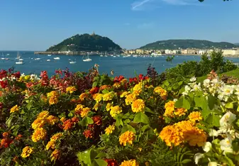Bright, colorful flowers in the foreground with a scenic view of San Sebastián’s coastline, boats in the harbor, and the iconic Monte Urgull rising in the background.