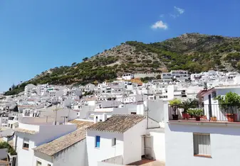 Cluster of traditional whitewashed houses climbing the hillside in Mijas, Spain, set against a backdrop of pine-covered mountains and a clear blue sky.