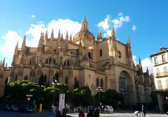 The grand Gothic-style Cathedral of Segovia with ornate spires and intricate stonework, bathed in golden light beneath a bright blue sky with puffy clouds.