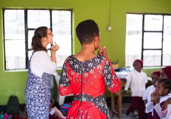 A volunteer teacher and a local student stand before a classroom of young children in Tanzania. They are pointing to their mouths to demonstrate pronunciation against a backdrop of bright green walls.