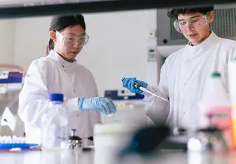 Two teenage students wearing lab coats, gloves, and safety goggles carry out a hands-on biomedical science experiment using a pipette in a modern laboratory setting.