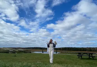 A traveler in overalls holds a mug and a pair of gloves while standing in a grassy coastal field at Seguinland, with a marsh and tree line in the distance