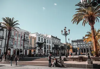 A wide-angle view of a historic town square with colonial buildings, palm trees, antique lamp posts, and pedestrians under a clear sky