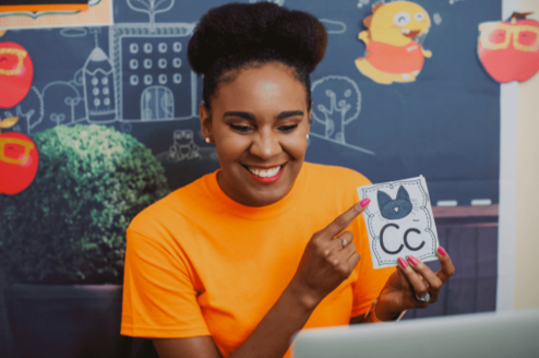 a woman in an orange shirt is smiling at a computer and pointing to a flashcard, which shows a picture of a cat and the letter C