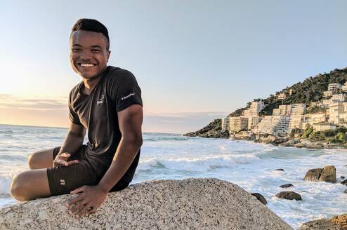 Alfred sitting on a boulder at the beach in South Africa smiling