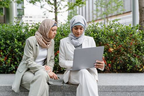 Two women with headscarves look at a computer screen outside.