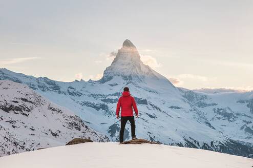 A person with a red puffer jacket stands on a summit facing The Matterhorn mountain in the alps