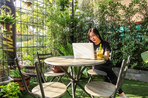 A woman looks at her laptop while sitting at a table surrounded by green plants.