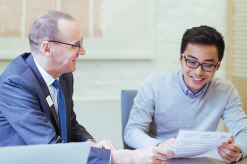 A male professor sits at a table next to a male student