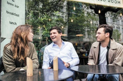 One female and two male postgraduate students sit at a table outside a cafe