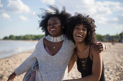 Two women with their arms around each other smile on a beach.