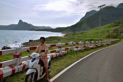 A man sits on a scooter in front of a bay and mountains.