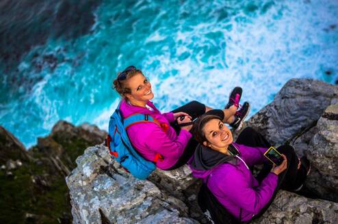 Two women sitting on a cliff by the ocean look up at and smile for the camera.