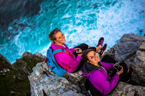 Two women sitting on a cliff by the ocean look up at and smile for the camera.