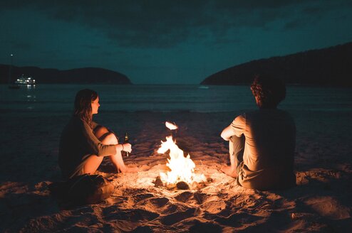 Two people sit by a camp fire on a beach at night.