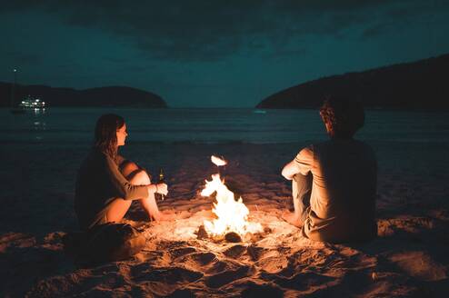 Two people sit by a camp fire on a beach at night.