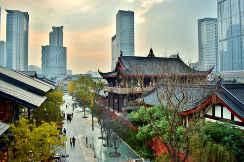 An image of Chengdu is shown. In the background, large city buildings can be seen, and in the foreground there are traditional Chinese buildings. On the left side of the image is a street lines with trees with people walking along it.