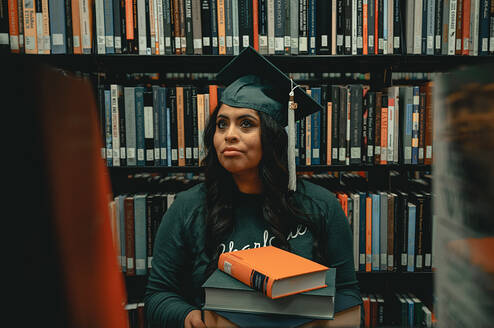 Student sitting next to bookshelf wearing graduation cap