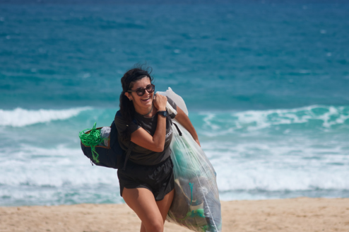 Woman walks along a beach smiling, carrying a bag of trash.