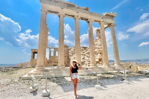 A young woman with long brown hair, wearing a black tank top and light denim shorts with a straw hat, stands in front of temple ruins with columns still standing.