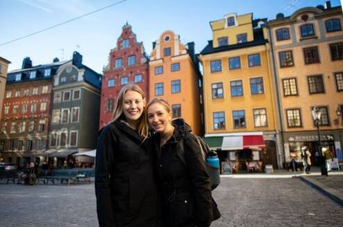 Two female students are standing in an old town square with colorful historical buildings behind them.