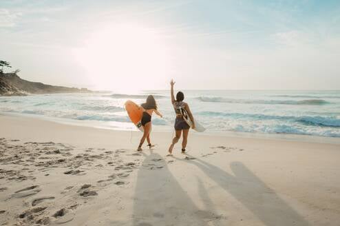 Two women holding surf boards walk on the beach.