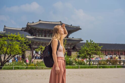A woman wearing a backpack shields her eyes from the sun with a temple in the background.