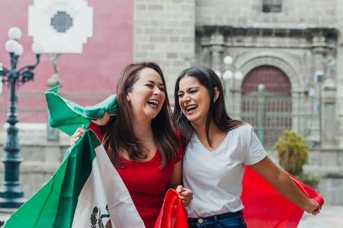 Two women laugh with a Mexican flag draped over their shoulders.