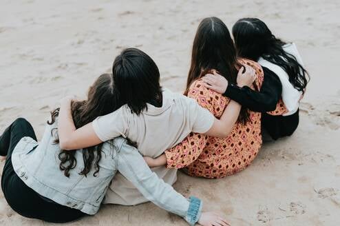 Friends with their arms around each other sit on the beach.
