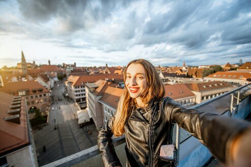 A woman smiles and takes a selfie on a rooftop with a German city in the background.