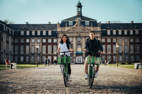 A man a woman ride bikes with a large building in the background.