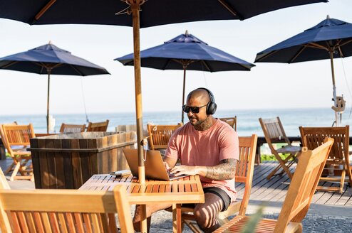A man sitting outside under an umbrella works on a laptop.