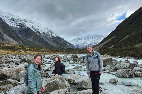 Three young women with backpacks stand on rocks and smile with mountains in the background.