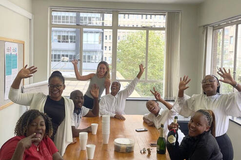 A group of people gathered around a table wave at the camera