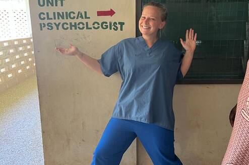 A volunteer in scrubs poses happily in front of a sign that reads 'Counselling Unit' and 'Clinical Psychologist'