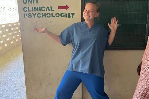 A volunteer in scrubs poses happily in front of a sign that reads 'Counselling Unit' and 'Clinical Psychologist'