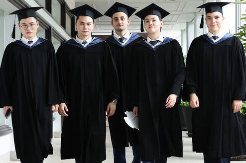 Five graduates in caps and gowns walk together down a hallway, facing forward and looking at the camera.