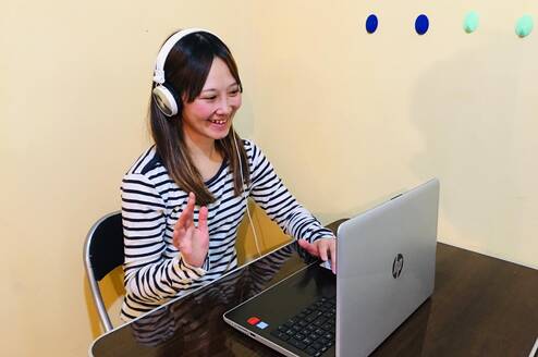 A Japanese teacher conducting an online 1-on-1 lesson with a student via Zoom. The teacher is smiling and explaining a lesson in Japanese using teaching materials on the screen.