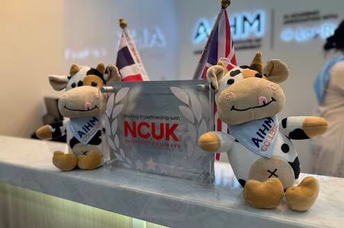 Two plush cow toys wearing AIHM College sashes sit on a reception desk next to a sign that reads, Working in partnership with NCUK University Pathways, with flags and logos in the background.