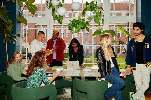 A group of students standing around a long table in front of a large white-pane window. The students are looking through text books, working on laptops, drinking coffee and chatting and laughing with one another. 