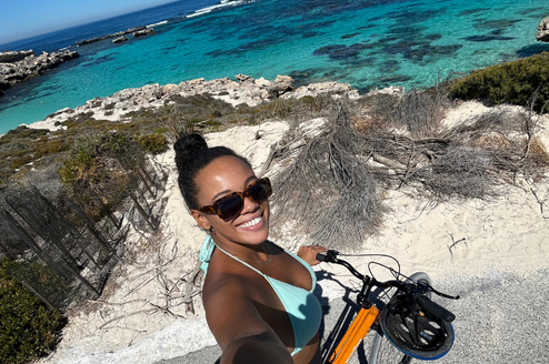 A girl on a bike taking a selfie with a blue sea behind her.