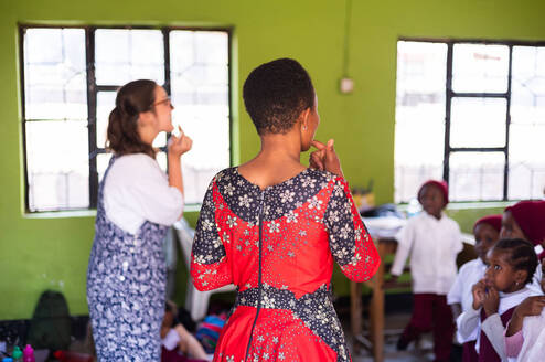 A volunteer teacher and a local student stand before a classroom of young children in Tanzania. They are pointing to their mouths to demonstrate pronunciation against a backdrop of bright green walls.