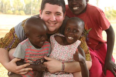 Volunteer kneeling outdoors with three young children, smiling and embracing them during a community program at Maisha.