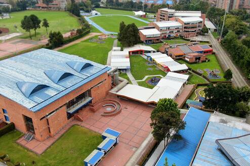 The image shows an aerial view of an educational institution with a spacious and well-organized campus, featuring brick buildings with blue and white roofs, well-maintained green areas, recreational spaces, and sports facilities such as an athletic track. Walkways and courtyards connect the different areas, and the surroundings combine nature with urban buildings, conveying a sense of order, openness, and institutional well-being.
