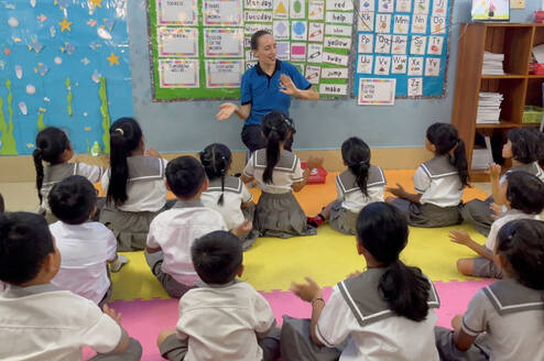 TEFL teacher sitting at the front of a classroom, engaging a group of young students in a lesson with hand gestures and visual aids on the wall.