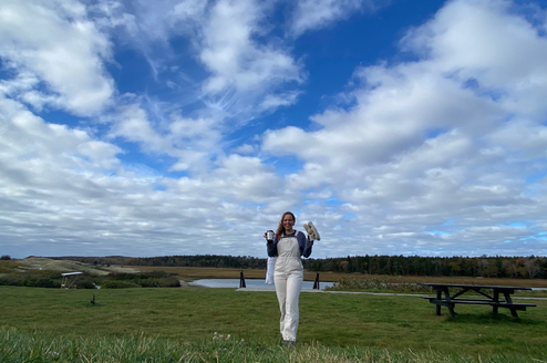 A traveler in overalls holds a mug and a pair of gloves while standing in a grassy coastal field at Seguinland, with a marsh and tree line in the distance