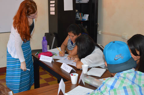 A woman leaning over a table while seated learners complete worksheets.