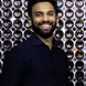 Vinay Vimalan wearing a navy button up shirt and a beard smiling