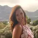Tamsyn in a pink and red floral dress ,with red long hair smiling, and mountains in the background 