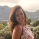 Tamsyn in a pink and red floral dress ,with red long hair smiling, and mountains in the background 
